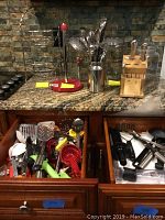 Kitchen counter setup showing two-tier fruit stand, three glass canisters, knife block with knives, cooking utensils in holder, and kitchen utensil drawers opened with assorted tools.