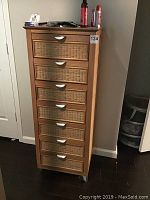 Front view showing all seven drawers with rattan woven fronts and silver handles, atop wheeled base.