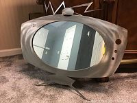 Full front view of oval metal mirror with space age TV style frame, pointed legs base and antenna top.