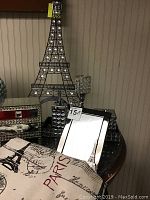 Black metal Eiffel Tower sculpture decorated with clear rhinestones, mirrored three-drawer box with crystal knobs, and black-bordered photo frame showing Eiffel Tower print on round table.