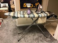 Photo showing folding ironing board with striped cover, Shark iron, and handheld steamer.