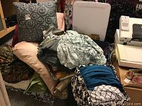 Overall collection of assorted linens, pillows, and bedspread stacked with storage bin and some shoes nearby