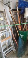 Photo showing a 5 foot aluminum ladder leaning against a wall next to garden tools and rakes with wooden handles and a green storage container.