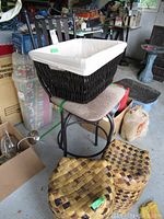 Photo of black metal swivel bar stool with beige upholstered seat and two palm leaf patterned baskets on the floor in front with a large black wicker basket with white liner on the stool.