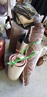 Photo showing multiple rolled carpet pieces and runners, some tied with green straps and a black plastic bag covering one, highlighting the variety of sizes and patterns.