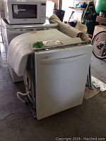 White Kenmore dishwasher shown with front partially covered by packaging material, placed on floor near other appliances.