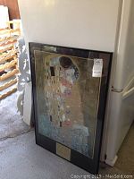 Framed poster leaning against a wall in a storage area, showing the entire artwork and frame.