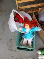 Cabbage Patch Kids doll in blue dress lying on a green plastic container next to a white bag filled with artificial red flowers