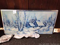 Full view of the winter scene painting with snow-covered houses and trees in a metal frame, placed on the ground against a wooden wall.