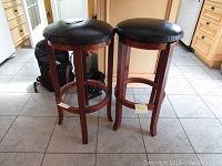 Pair of black leatherette cushioned kitchen bar stools standing side by side on a tiled floor, showing the wooden frame and cushion shape.