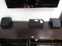 Two satellite speakers and one mini center speaker placed under a Samsung flat screen TV on a wood surface.