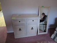 White hall stand cabinet positioned next to rectangular white framed wall mirror, showing front and side angles.