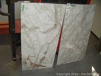 Two white marble slabs leaning against a black metal frame and a red background, showing veining and a brown stain on one slab near the bottom corner.