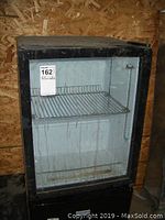 Front view of a black Avanti wine fridge with glass door and single wire shelf inside showing dust and needing cleaning.