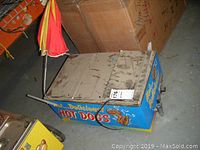 Front-left perspective showing blue cart body, vinyl “HOT DOGS” graphics, hinged lids and one wheel.