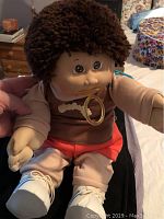Front view of the Cabbage Patch doll showing face, curly brown hair, brown shirt with bone applique, and translucent ring held by doll.