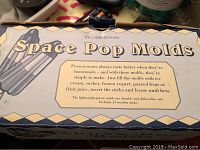 Box front showing Williams Sonoma Space Pop Molds title and details about the product including making frozen treats with various fillings, highlighting lightweight plastic molds, dishwasher safe, and inclusion of 24 wooden sticks.