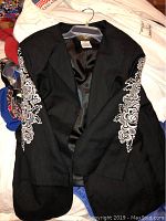 Front view of black Bob Mackie jacket with detailed white floral embroidery on the sleeves, displayed on hanger.