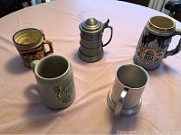 Five assorted beer steins shown on a table, including metal and ceramic styles