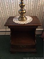 Photo of one Ethan Allen solid wood nightstand, showing the top surface with a brass lamp base on a lace doily, drawer with brass handle and lower shelf