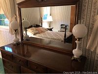 Wide view showing both vintage milk glass globe lamps positioned on a wooden dresser in front of a large mirror in a bedroom.