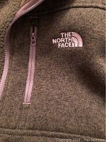 Close-up image of the chest area showing The North Face white embroidered logo and chest pocket zipper on gray fleece material