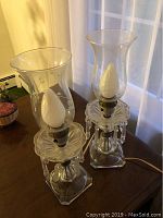 Pair of matching glass hurricane lamps on a wooden surface near a window. Clear glass with decorative hanging prisms and etched designs on the hurricane shades.