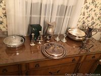 Thirteen silver and pewter pieces arranged on a wooden sideboard including candlesticks, trays, bowls, and covered dish.