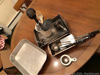 Photo showing metal baking pans, measuring cups, and a box container holding various utensils including metal ladles and spatulas on wooden table.