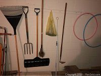 Photo showing garden tools hung on a wall including a large leaf rake, two pitchforks, a snow shovel, a metal stirrer, a hole digger, and three colorful hula hoops on the wall.