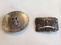Two metal belt buckles shown side by side on white background. Left is oval with raised skull and circular border indentations, right is rectangular with detailed eagle or phoenix and flames design.