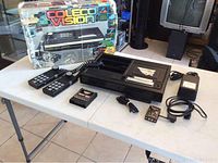 Wide view showing the ColecoVision gaming system with two numeric keypad controllers, Donkey Kong cartridge, power adapter, cables, and original box with wear and damage.