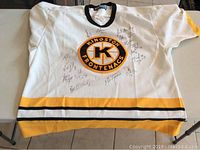 Front view of white Kingston Frontenacs hockey jersey with signatures on front around team logo, showing black and yellow trim