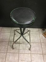 Front angle showing the metal stool with black cushioned seat with green paint on seat edges and sturdy metal legs with slight rust.