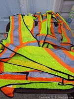 Three fluorescent yellow-green safety vests with bright orange borders and grey reflective ScotchLite strips, laid out to show front and back details, including Velcro adjustment and pockets.