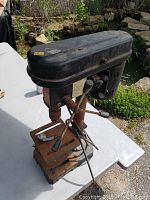 Side angled view of the electric drill press showing the black motor housing, rusted metal base, and operational handles.