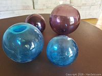 Four handblown glass balls in blue and purple, two larger and two smaller, arranged on a dark wooden surface.
