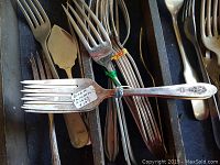 Close-up of various forks and spoons with one fork showing a paper tag with details: '31 pc set Brazil Silver 6 1/2" 12/7'