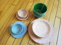 Powder blue cup with matching saucer; blush petal cup with matching saucer; stacks of green bowls and blush petal dinner and side plates arranged on wooden floor.