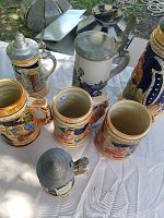 Photo showing multiple German beer steins of varying sizes and designs on a white surface outdoors, highlighting the colorful painted scenes and pewter lids.