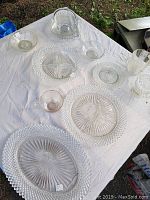 Top-down photo showing 10 clear textured glass items on a white cloth: large oval platter, round plates, bowls, creamer/pitcher, and glasses arranged closely together