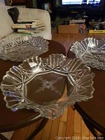 Three clear glass bowls with intricate scalloped edges and patterned surfaces, displayed on two small round tables in a living room setting.