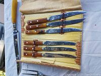 Seven knives and one carving fork with yellowish horn-like handles inside a yellow and brown leather-like case