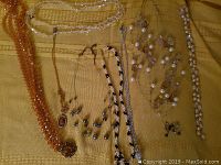 Full view of 8 necklaces arranged on yellow cloth, varying in bead color and style, including chains and pearl-like beads.