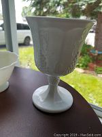 White milk glass chalice with embossed grapevine pattern on a pedestal base.
