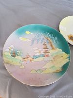 Hand painted decorative plate showing detailed Asian landscape with pagodas, trees, and a bridge, pastel colors with gold accents.