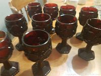 Set of ten ruby glass goblets arranged on a wooden table, showing their deep red color, hexagonal bases and decorative patterning.