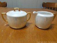 Photo showing the white porcelain sugar dish with lid, and matching creamer with gold handles, on a wooden table.