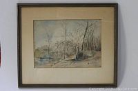Framed watercolor landscape painting showing bare trees and a body of water in subtle earth tones.