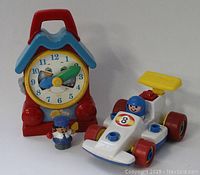 Photo showing vintage Fisher Price toy clock with red roof handle, blue frame, and clock face with character eyes and smiling mouth, alongside white and red race car toy with number 8 and blue-helmeted driver figure.